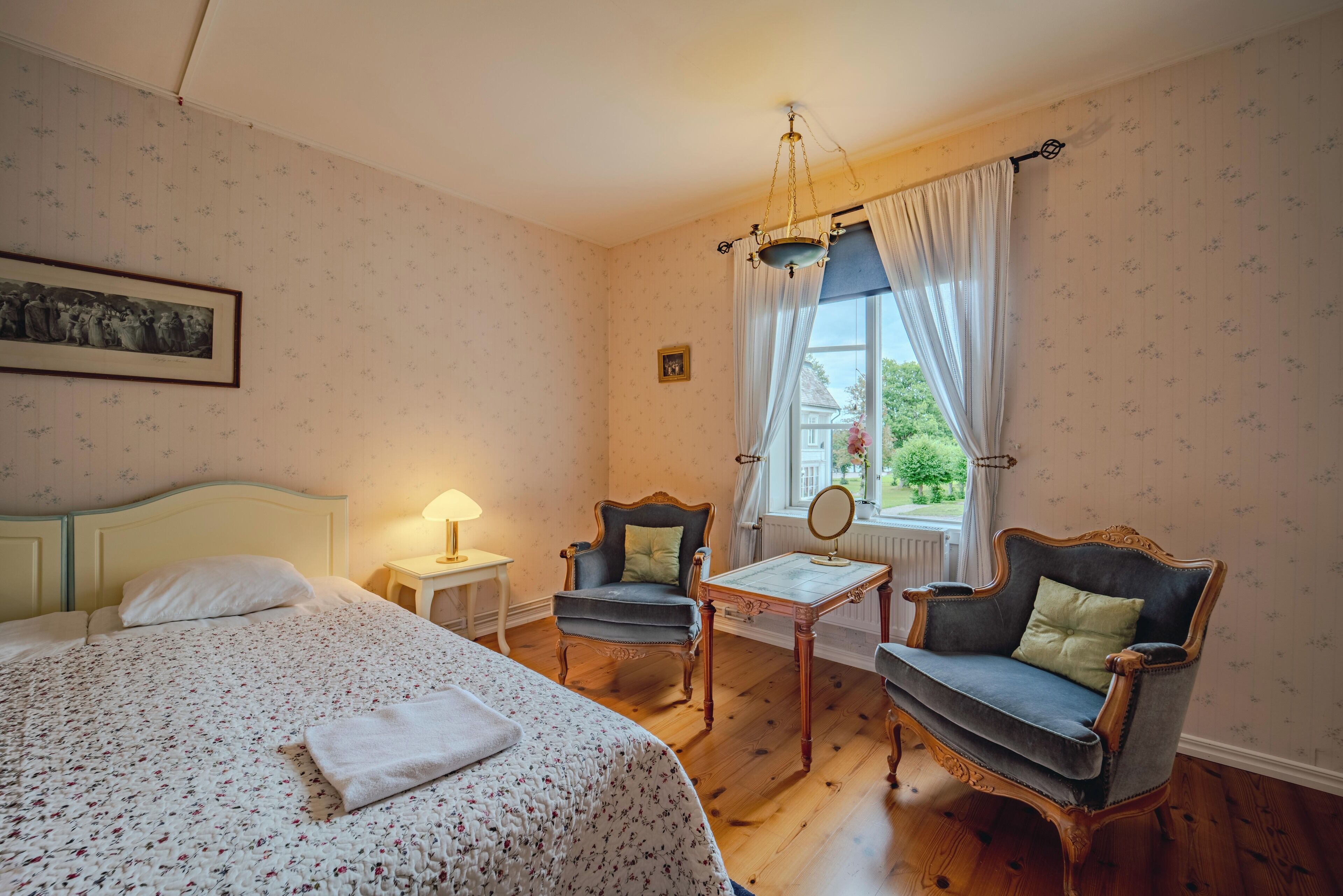 A bedroom with a bed, two armchairs, a small table, a lamp, and a window with curtains.