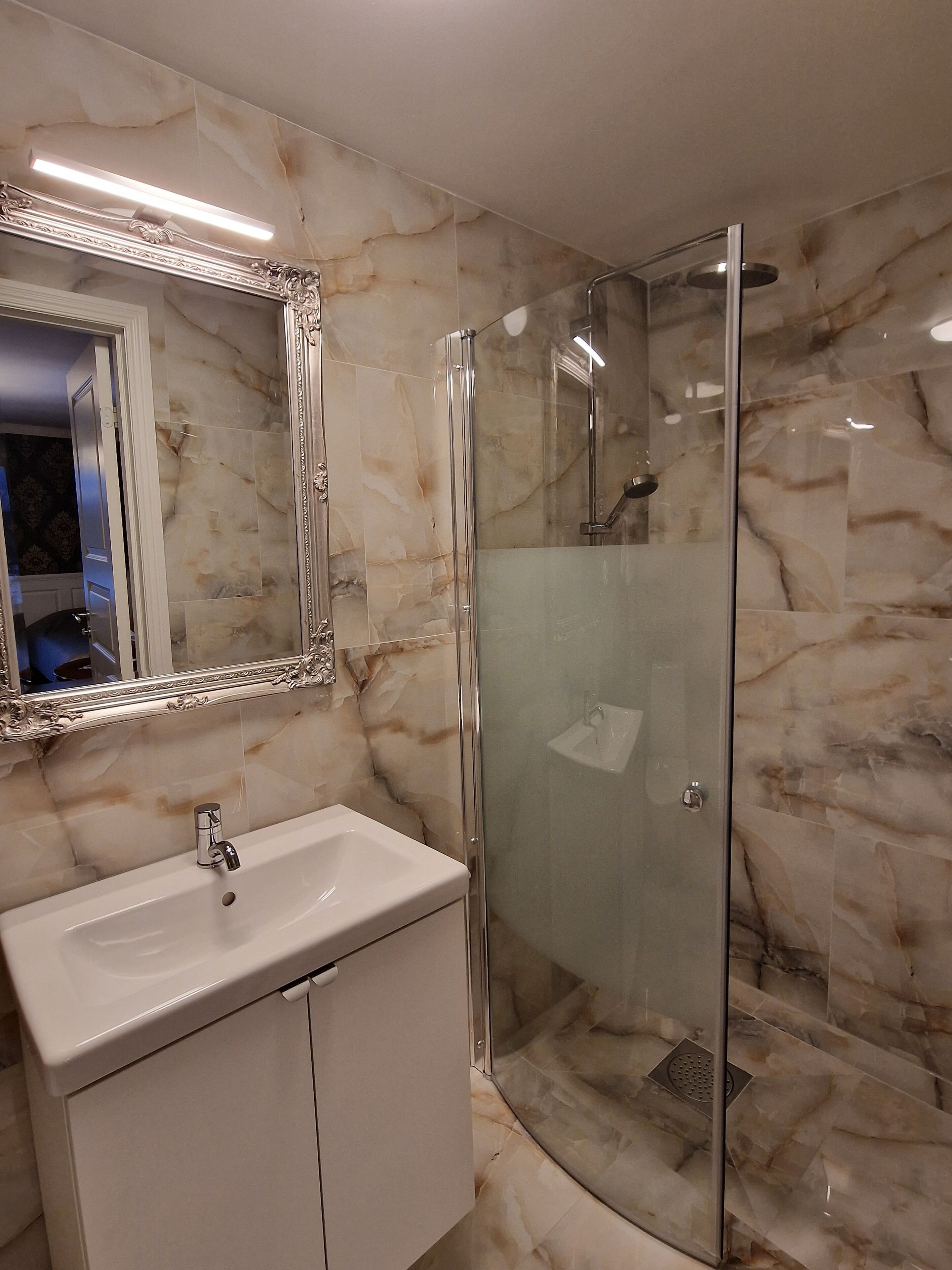 Une salle de bain avec un mur en marbre, une douche en verre, un lavabo blanc et un grand miroir.