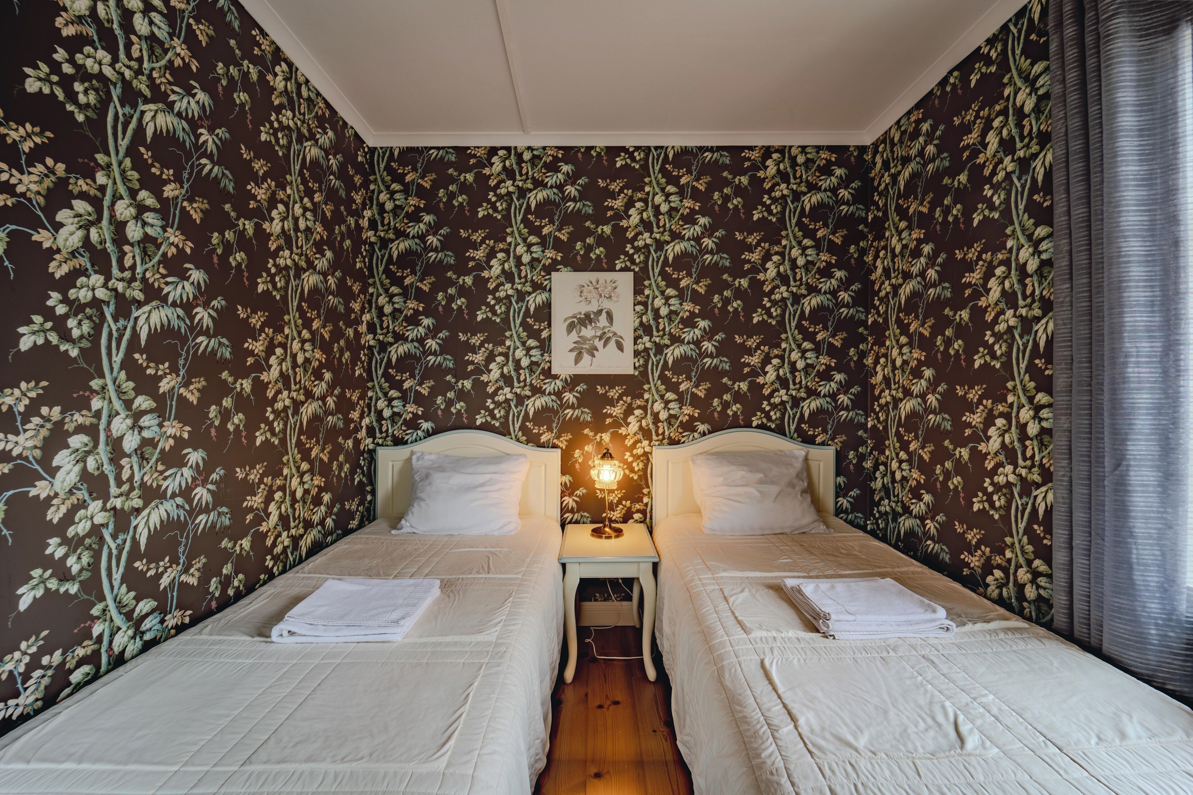 Ein Zimmer mit zwei Betten, einem Nachttisch mit Lampe und blumigem Wandpapier.