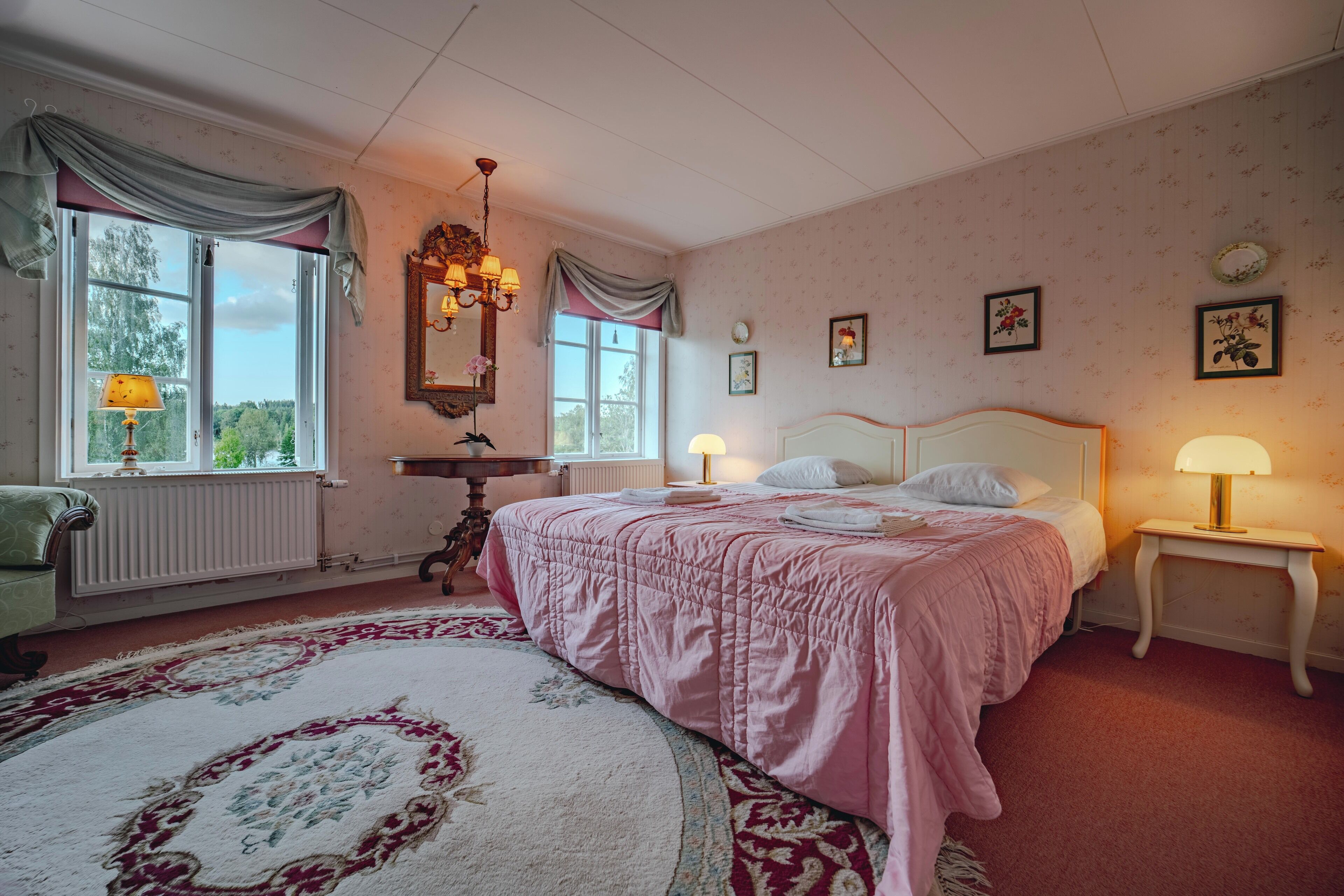 Um quarto com uma cama grande, uma mesinha de cabeceira, cômoda, janela com cortinas, lustre e radiador.