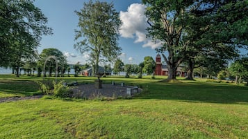 Ein grasbewachsenes Gelände mit einem großen Baum, einem Steinfbrunnen und einem roten Gebäude im Hintergrund.