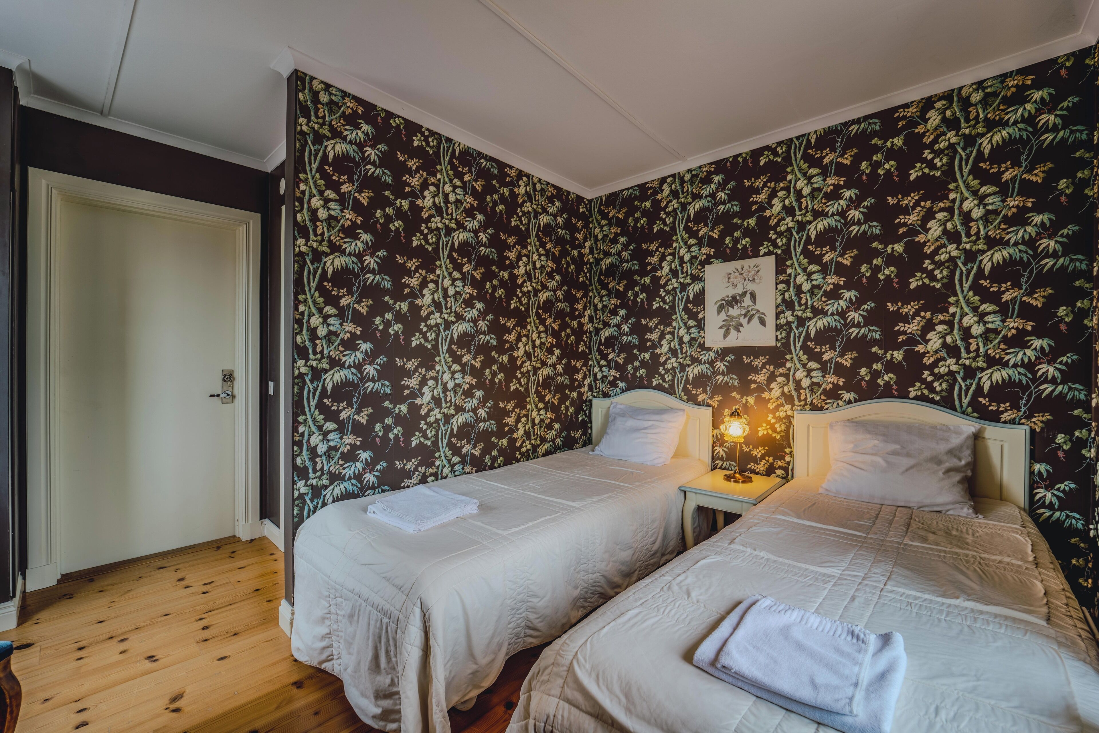 A room with two beds, wooden flooring, floral wallpaper, and a framed picture on the wall.