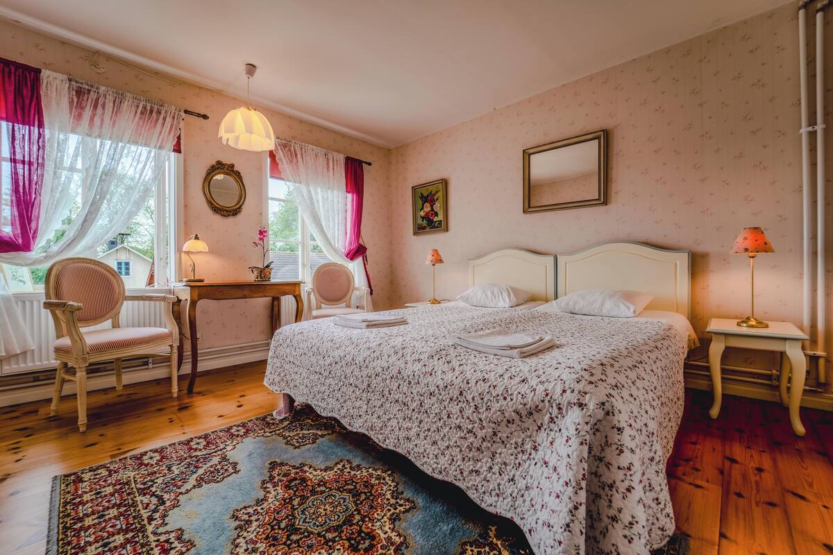 A bedroom with a large bed, two bedside tables, a chair, a desk, and a window with curtains.