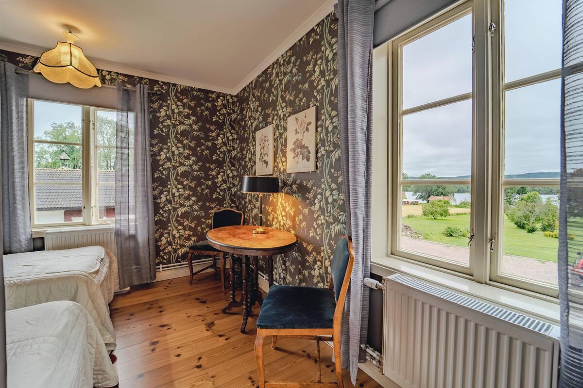 A room with a bed, a table with a chair, a window with a view of the outdoors, and a wall with floral wallpaper.