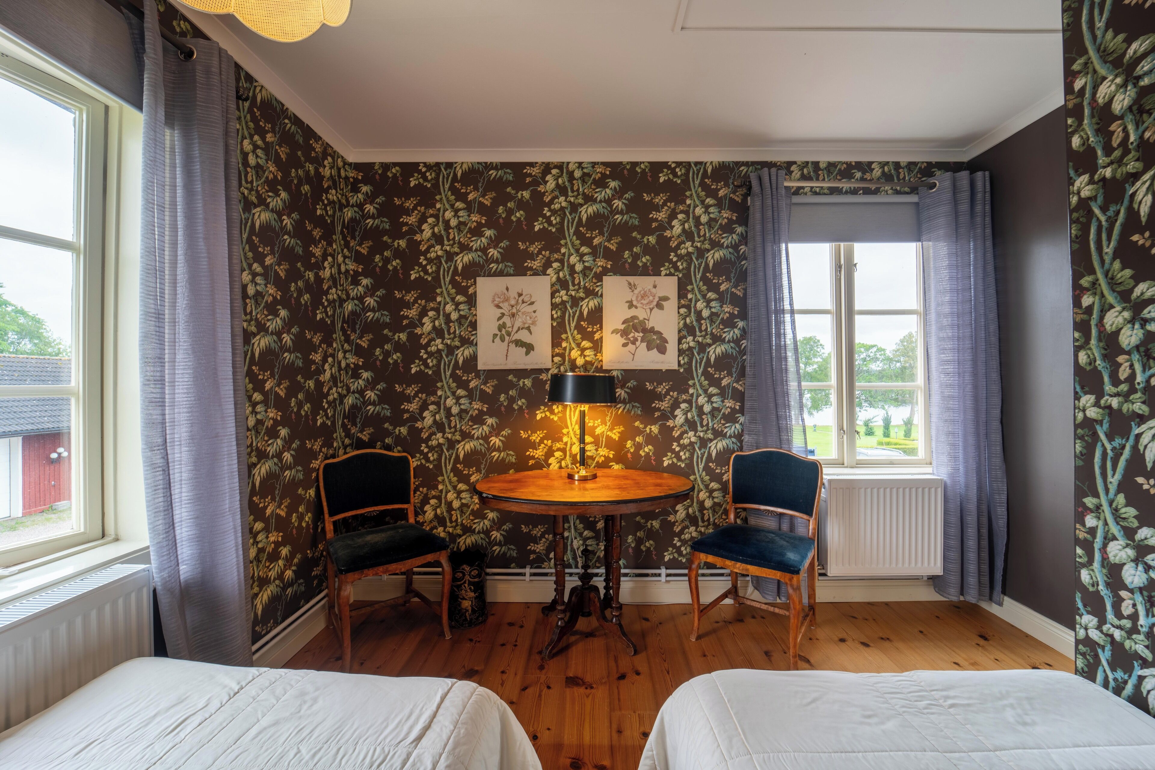 A room with two beds, a table with a lamp, two chairs, and floral wallpaper.