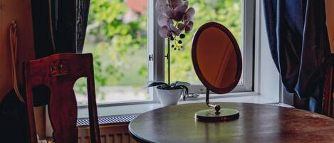 Una habitación con una mesa redonda de madera, dos sillas, una planta en maceta, un espejo y una ventana con cortinas.