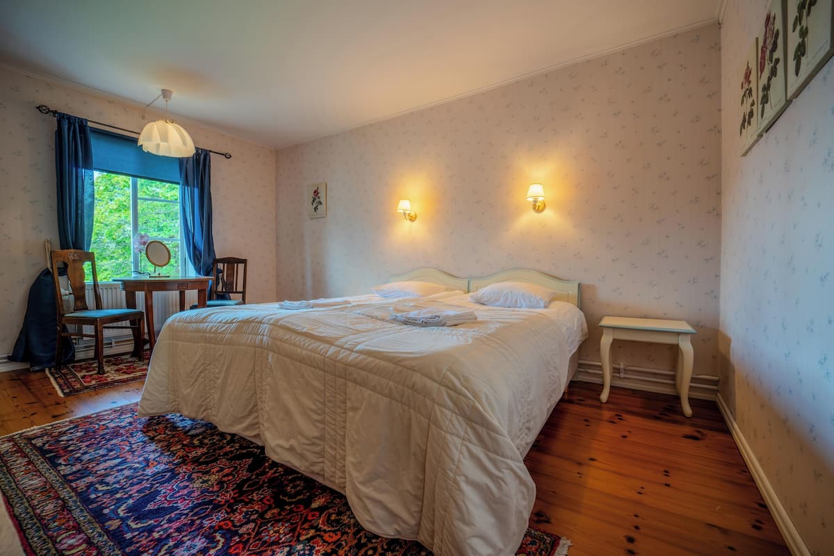 A bedroom with a large bed, two bedside tables, a chair, a desk, and a window with curtains.