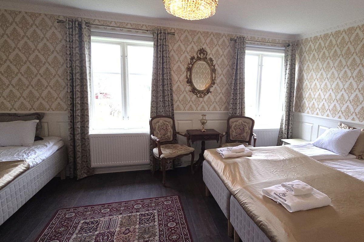 A hotel room with two single beds, a sitting area with a chair, a table, and a large mirror on the wall.
