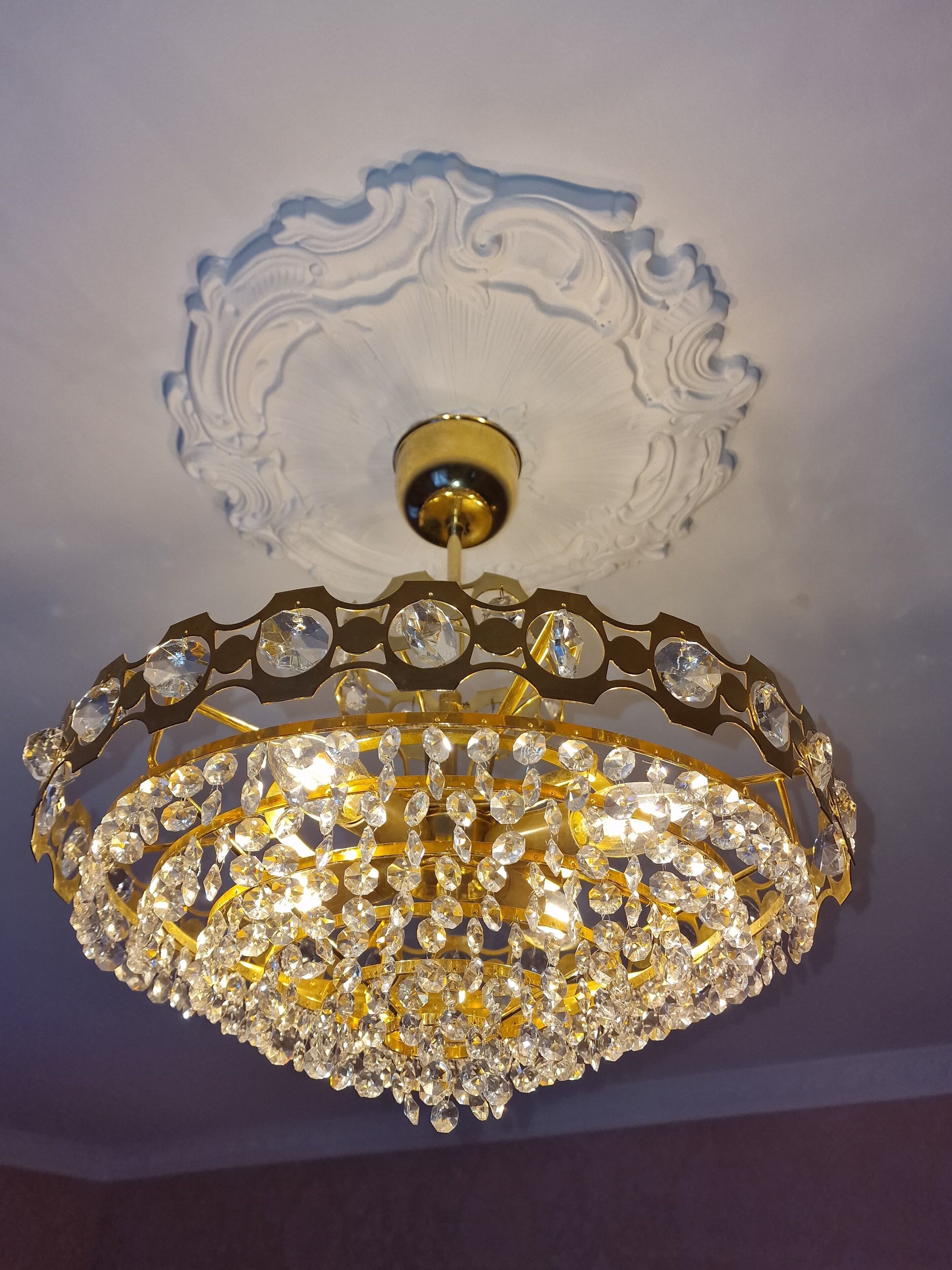 Ein Kronleuchter mit zahlreichen Kristallapplikationen und goldener Rahmen, der an einer kunstvoll gestalteten Decke hängt.