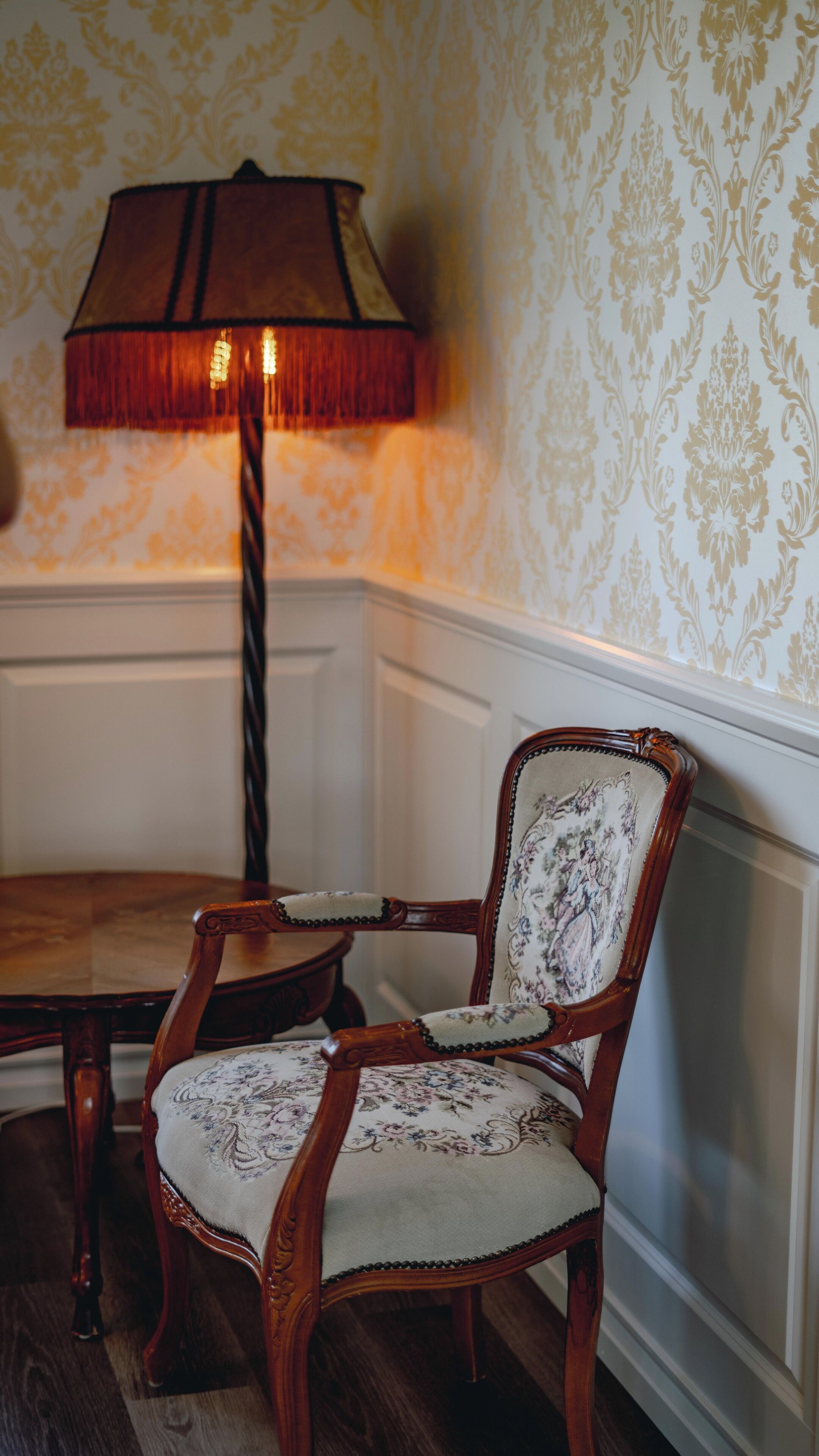 A wooden chair with a floral upholstery, a table, and a lamp with a red shade.