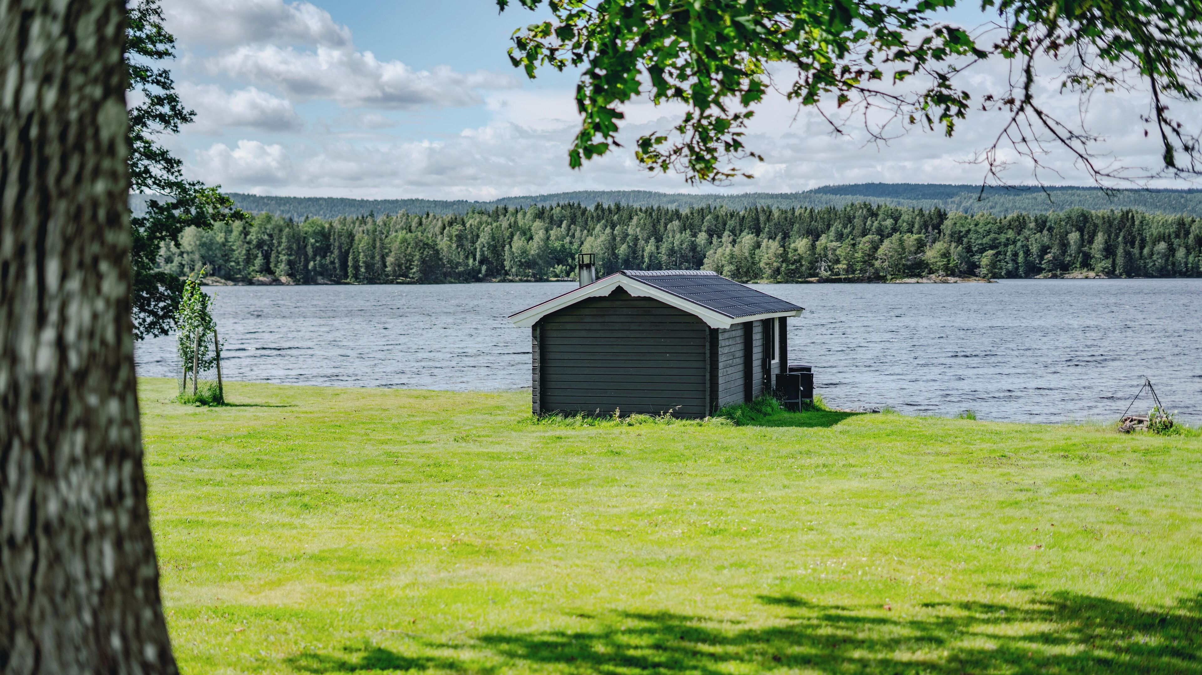 A small wooden cabin with a sloped roof, situated on a grassy area near a lake, surrounded by dense forest.
