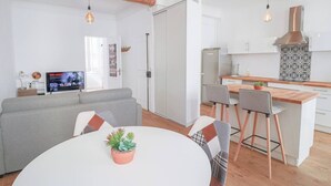 Una sala moderna con un sofá gris, una mesa de comedor blanca con una planta en maceta y una zona de cocina con encimera de madera y armarios blancos.