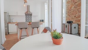 Una cocina moderna con armarios blancos, techo de madera y pared de piedra. Hay un comedor con una mesa y sillas blancas, y una planta en maceta sobre la mesa.