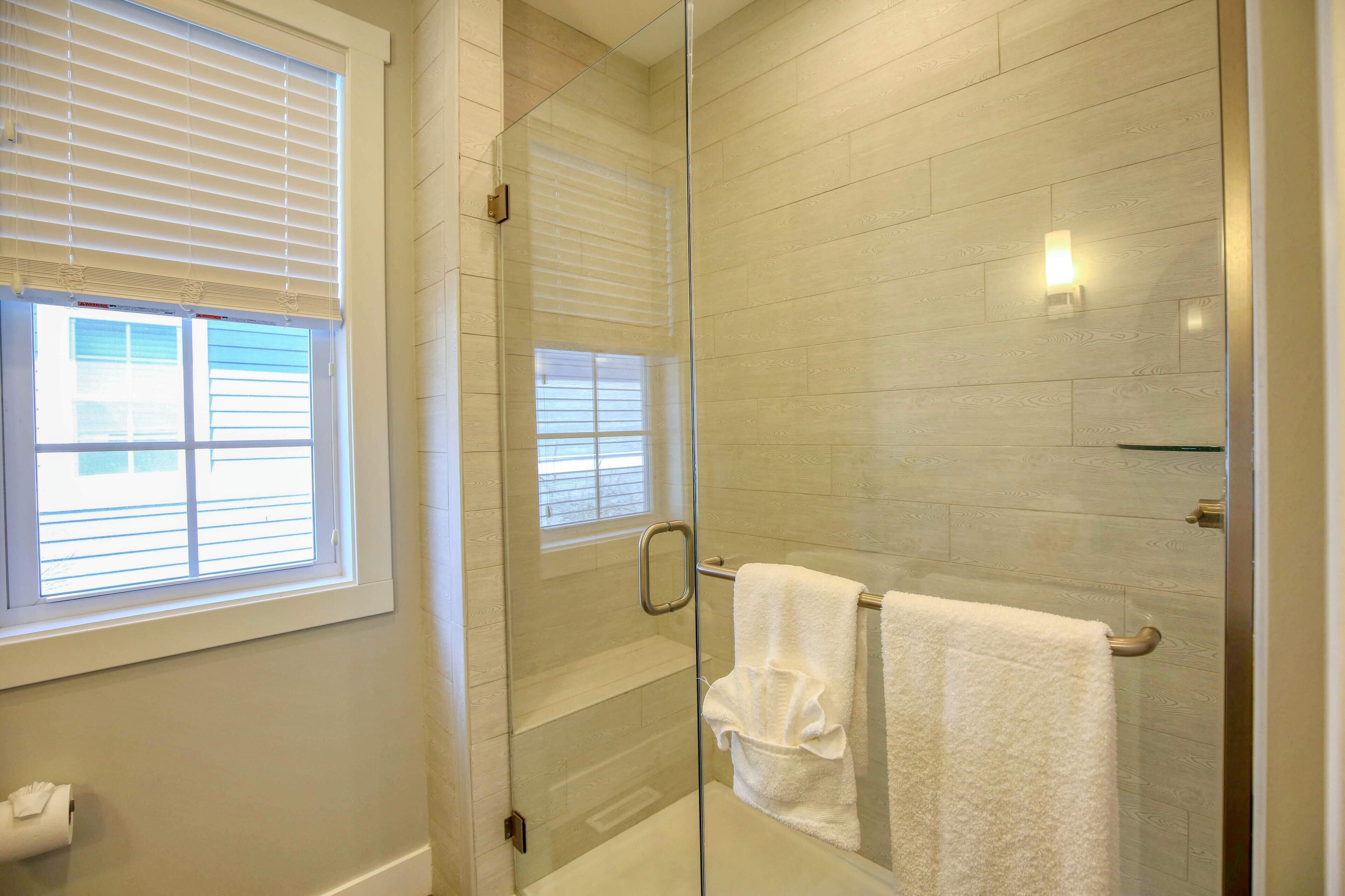 A glass-enclosed shower with a white towel rack, a window with blinds, and a light fixture on the wall.