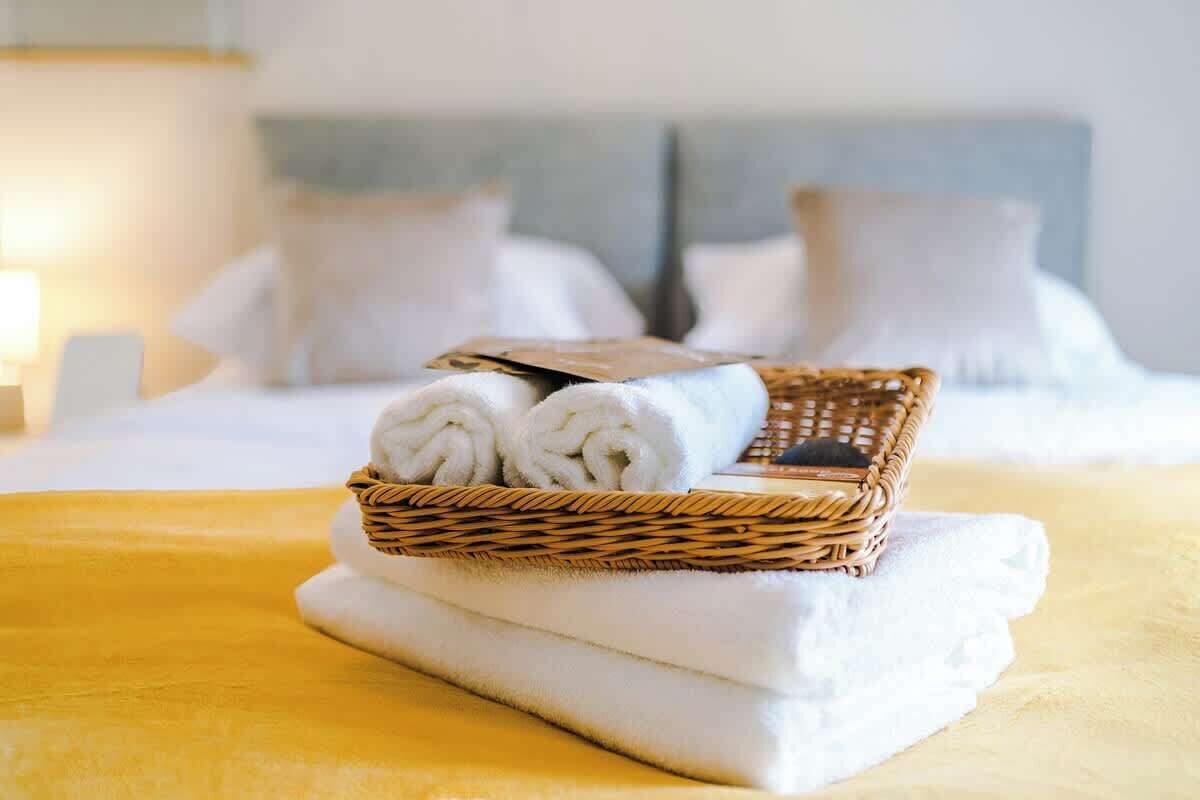 A neatly made bed with white linens, a wicker basket with rolled towels, and a folded towel on a yellow bedspread.
