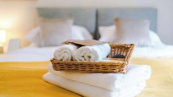 A neatly made bed with white linens, a wicker basket with rolled towels, and a folded towel on a yellow bedspread.