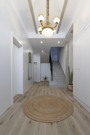 Een moderne hal met een houten vloer, een rond geweven vloerkleed, een kroonluchter, een spiegel, een trap en een cactusplant.