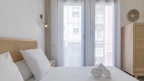 Une chambre d’hôtel avec un lit, des oreillers blancs, une serviette roulée, une tête de lit en bois, une lampe fixée au mur et une vue à travers des rideaux transparents.