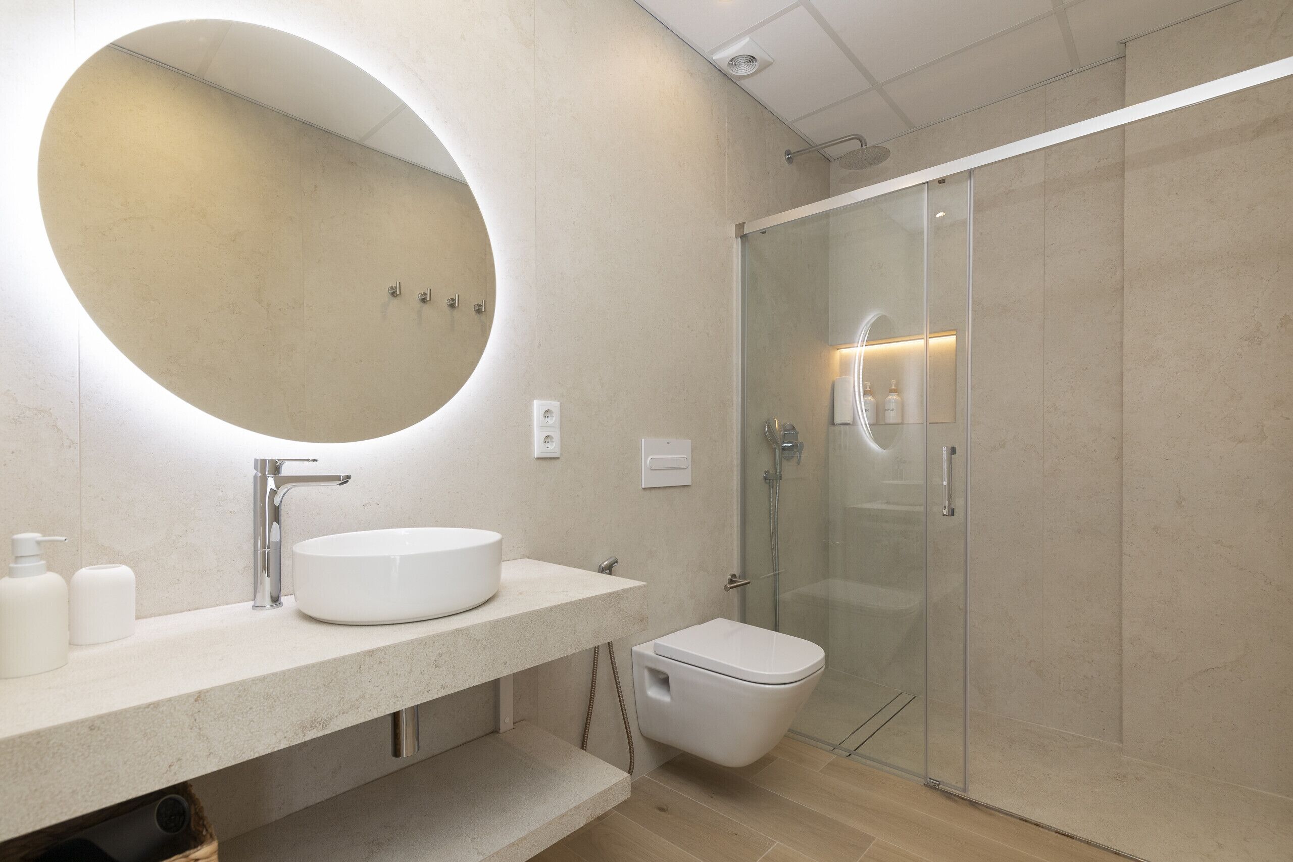 A modern bathroom with a round mirror, a white sink, a toilet, and a glass-enclosed shower.