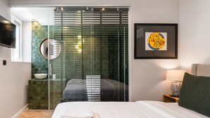 Una habitación de hotel moderna con una cama grande, un box de ducha de vidrio, un espejo redondo, una obra de arte enmarcada y una mesita de noche con lámpara.