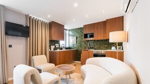 Una sala moderna con sofá, sillones y una mesa de centro. Una cocina con armarios de madera y salpicadero verde es adyacente.