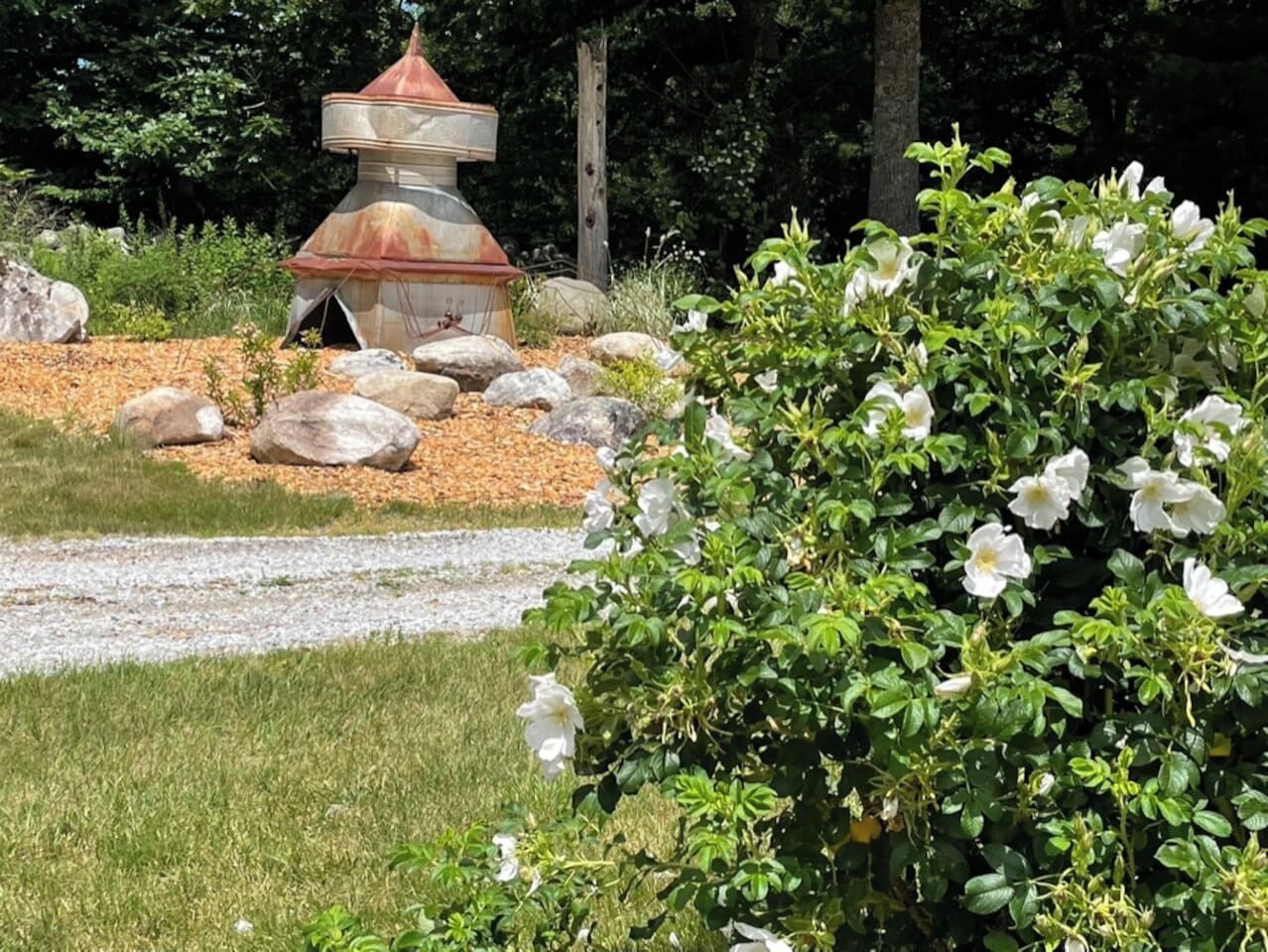 Un kiosque doté d’un toit conique, entouré d’un chemin de gravier, et un arbuste aux fleurs blanches au premier plan.