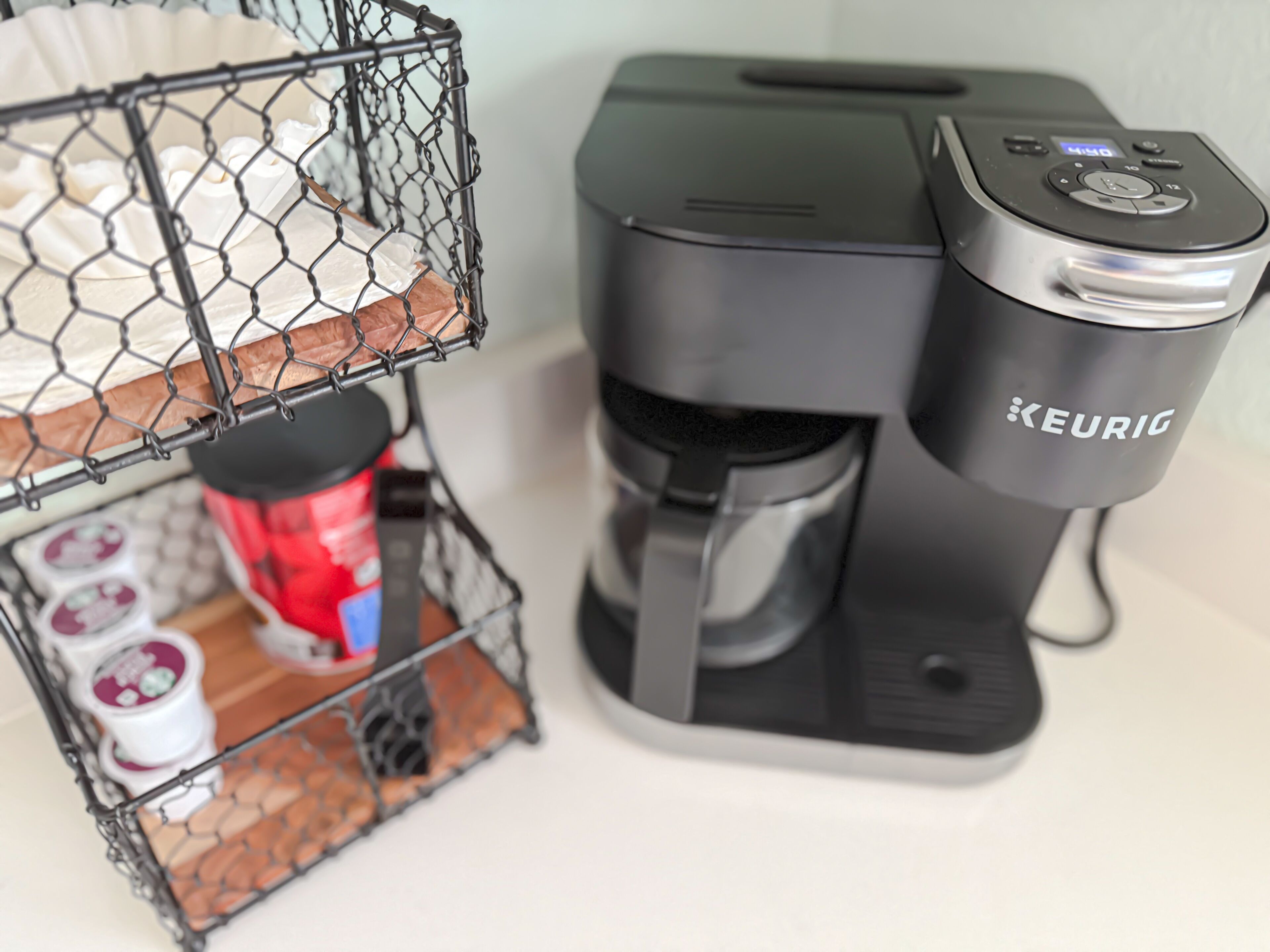 A Keurig coffee maker on a white countertop with a coffee pod holder and a basket containing a red coffee pod and two white pods.