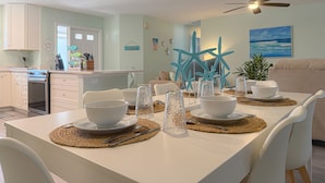 Un coin repas avec une table blanche dressée pour quatre personnes, des décorations en forme d’étoiles de mer et un ventilateur de plafond.