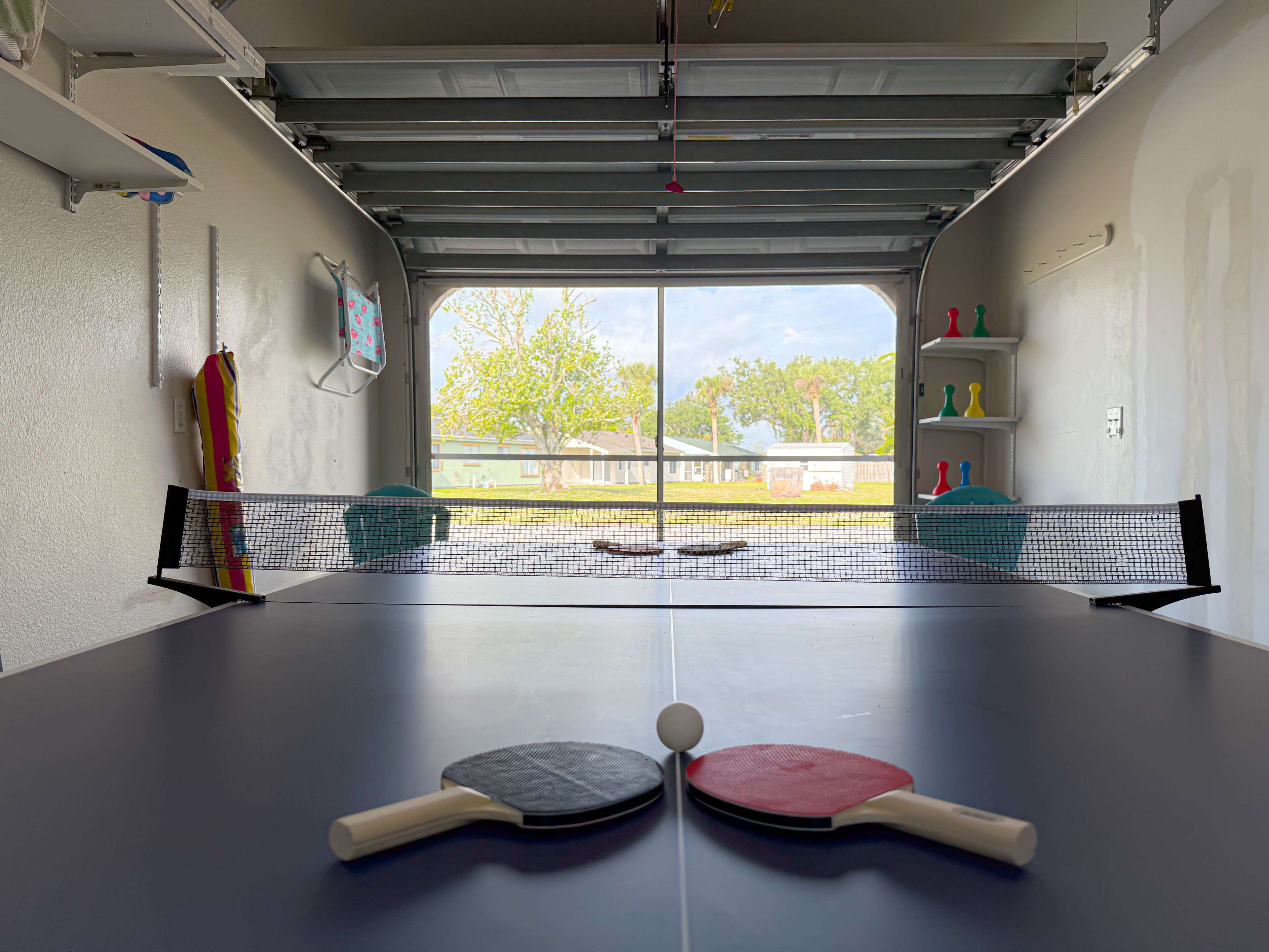 Una mesa de ping pong con dos palas y una pelota en una habitación con puerta de garaje y vistas a una zona residencial.