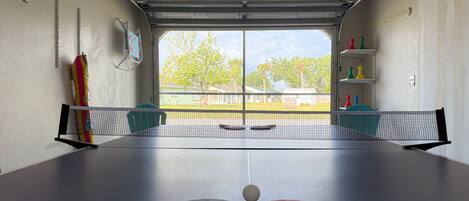Una mesa de ping pong con dos palas y una pelota en una habitación con puerta de garaje y vistas a una zona residencial.