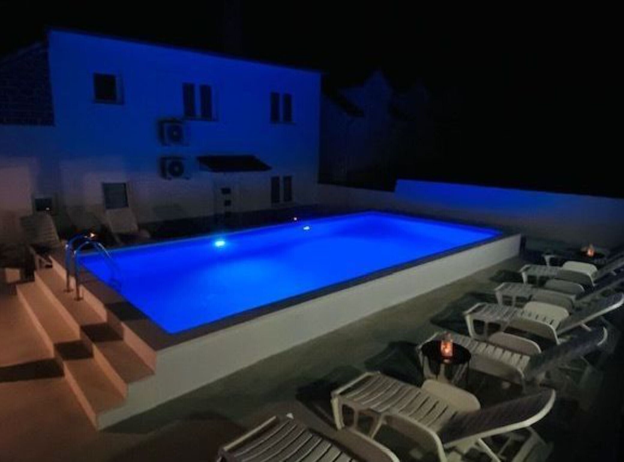 Una piscina con borde iluminado en azul, rodeada de sillones de descanso y un edificio de techo plano.