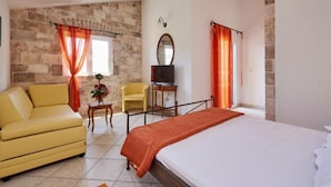 Quarto de hotel com cama, sofá amarelo, mesa pequena com um vaso, poltrona e televisão em um suporte.