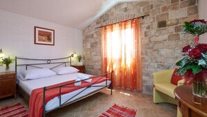 A bedroom with a stone wall, a bed with a metal frame, a wooden nightstand, a bedside table with a lamp, a red and white striped rug, a yellow armchair, and a vase with red roses. - Krknjasi - Double Bedroom (Drvenik Veliki)