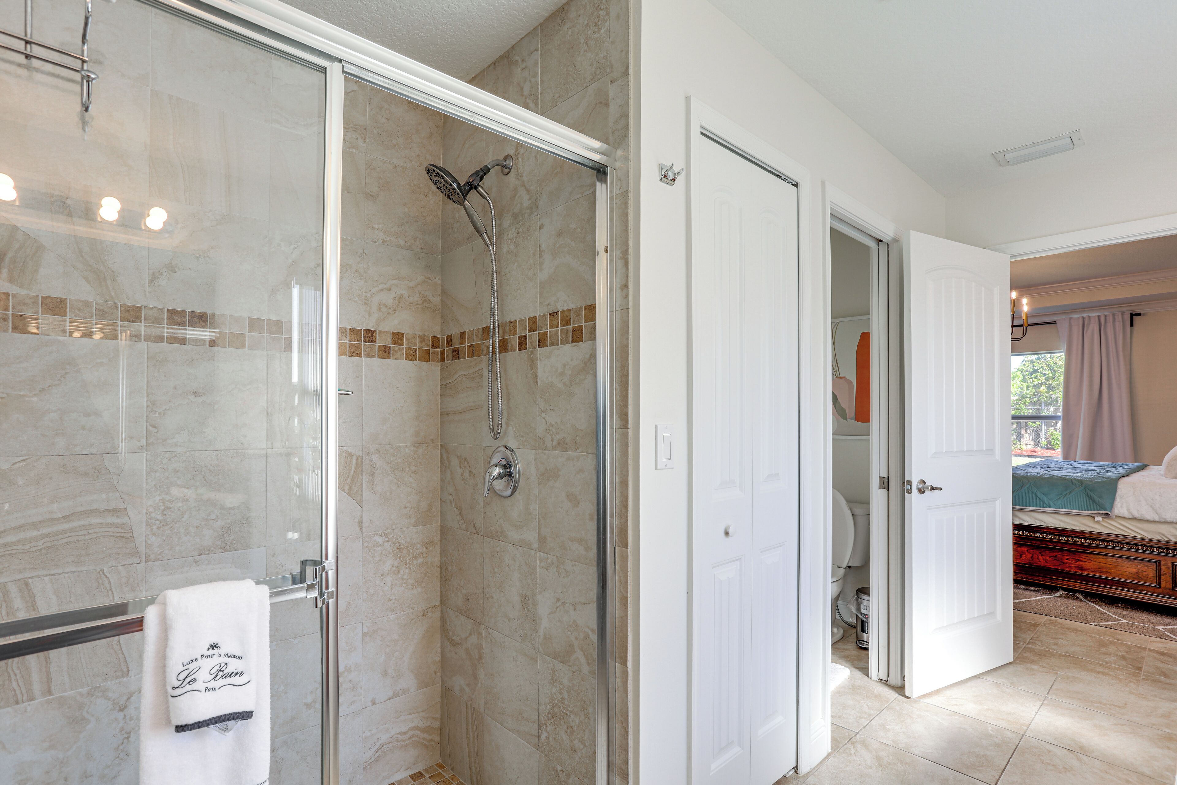 A modern bathroom with a glass-enclosed shower, a white towel hanging on a rack, and a visible door leading to a bedroom with a bed and a window.