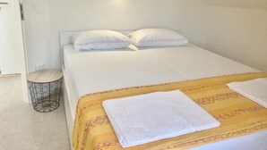 A single bed with a yellow patterned bedspread and white pillows, a round metal side table, and a small round trash bin. - One Bedroom Apartment with Balcony (Sutivan)
