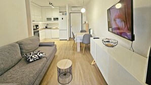 A modern living room with a grey sofa, a round coffee table, and a dining area with a table and chairs. A kitchen with white cabinets and appliances is visible in the background. - One Bedroom Apartment with Private Garden (Solin)