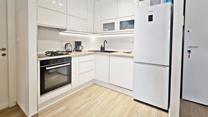 A modern kitchen with white cabinets, a built-in oven, a gas stove, a coffee maker, and a white refrigerator. - One Bedroom Apartment with Private Garden (Solin)