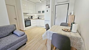 A modern kitchen with white cabinets, a stove, and a coffee maker. There is a dining area with a table covered in a lace tablecloth and two chairs. - One Bedroom Apartment with Private Garden (Solin)