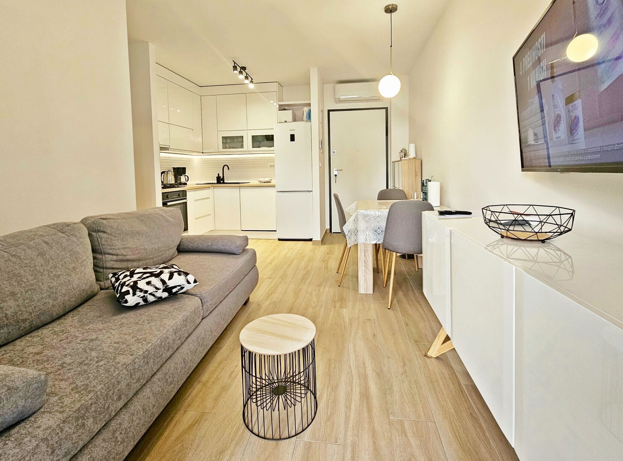 A modern apartment with a grey sofa, a round wooden side table, and a kitchen area with white cabinets and a dining table.