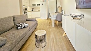 A modern apartment with a grey sofa, a round wooden side table, and a kitchen area with white cabinets and a dining table. - One Bedroom Apartment with Private Garden (Solin)