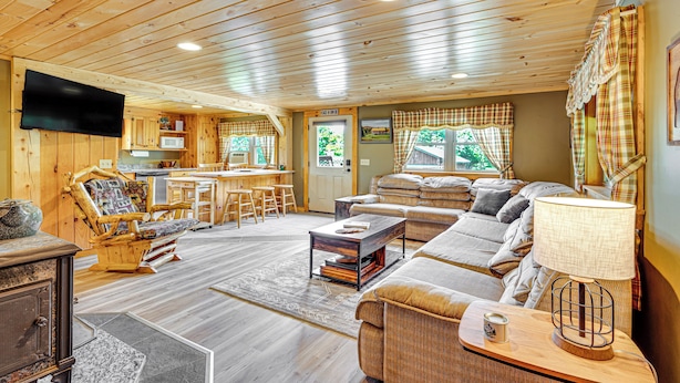 Ein gemütliches Blockhaus-Interieur mit Holzbalkendecke, einem Wohnbereich mit Sofa und Sesseln, einem Essbereich mit Tisch und Stühlen sowie einer Küche mit Mikrowelle und Fernseher.