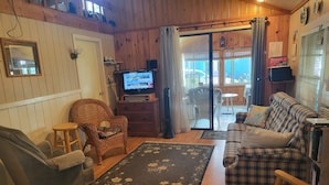 Un salon chaleureux avec un canapé, un fauteuil inclinable, une chaise en osier, un téléviseur posé sur un meuble en bois et une vue sur l’extérieur grâce à une porte vitrée.