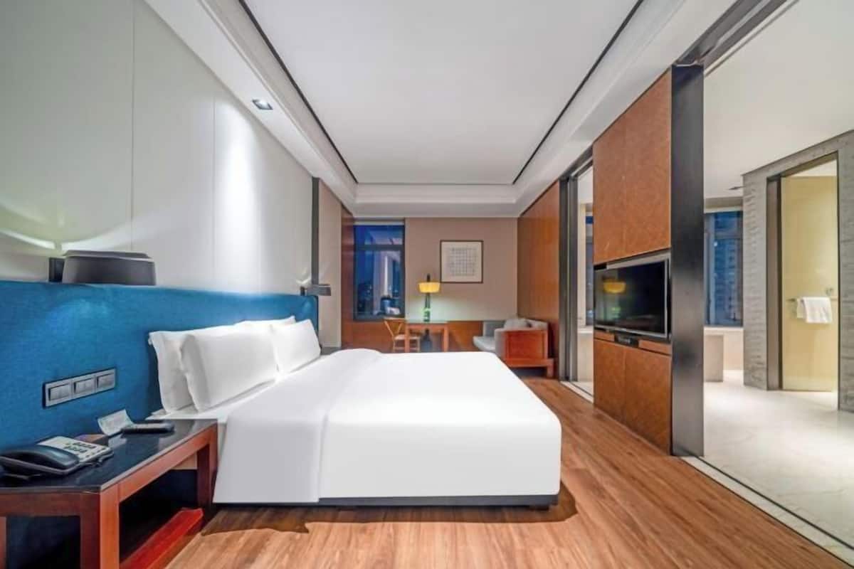 Habitación de hotel con cama, mesita de noche, lámpara, televisor y vistas al exterior.