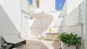 A small, sunlit outdoor area with a white wall, a white door, a white bench, a table with a white cushion, a white planter with green plants, and a white umbrella. - Residenza Vico Recupero by Wonderful Italy (Polignano a Mare)