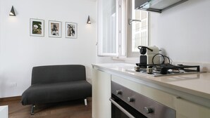 A modern kitchen with a built-in oven, a gas stove, and a window with blinds. There is a dark-colored sofa against the wall and three framed pictures on the wall. - Residenza Vico Recupero by Wonderful Italy (Polignano a Mare)