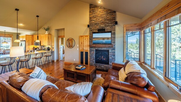 A spacious living area with a stone fireplace, leather sofas, and a large window offering a view of the outdoors. - Cabin 75 Brasada Ranch + Hot Tub and Golf (Powell Butte)