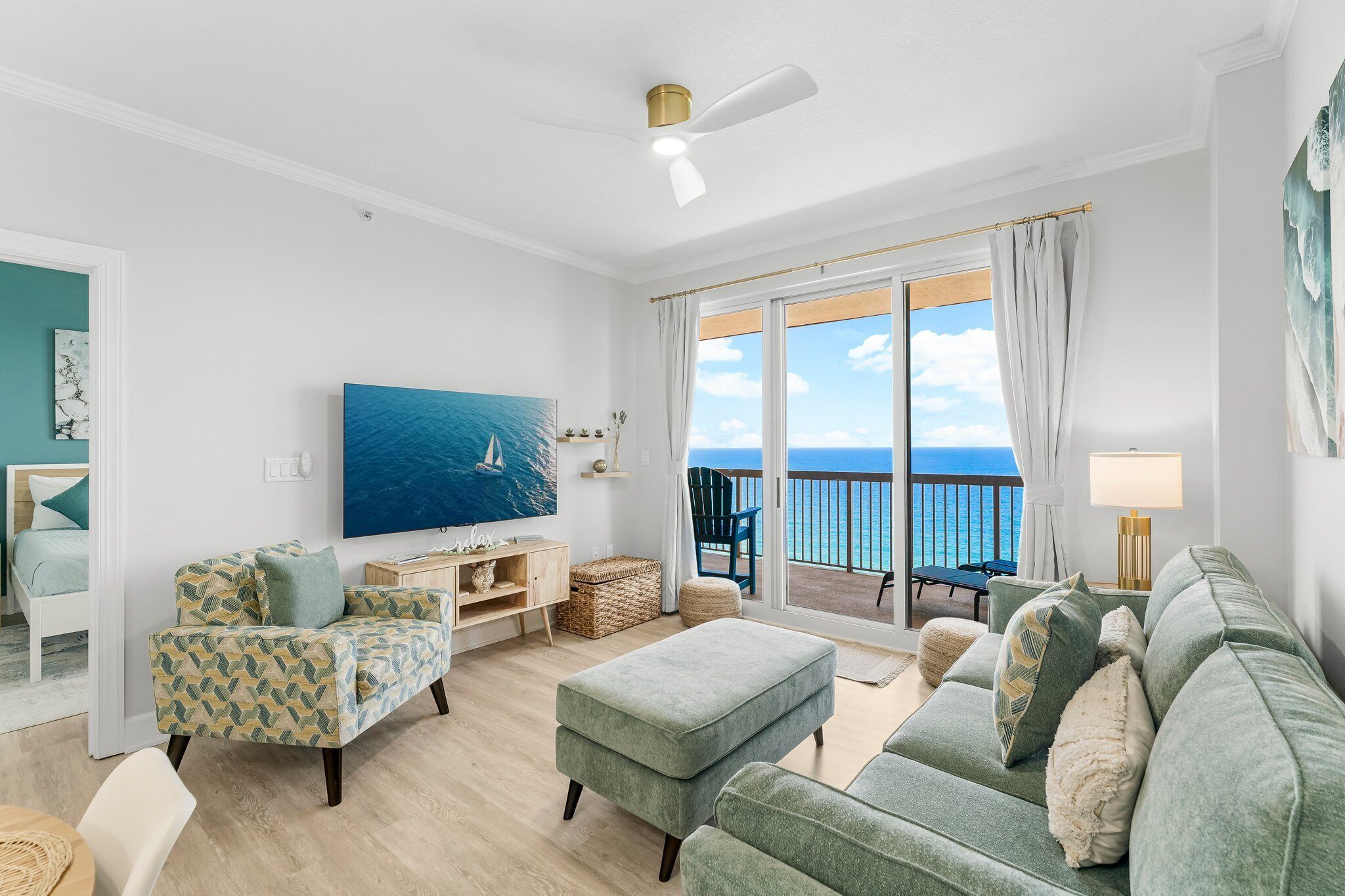A living room with a sofa, ottoman, and armchair, a television, and a view of the ocean through a sliding glass door.