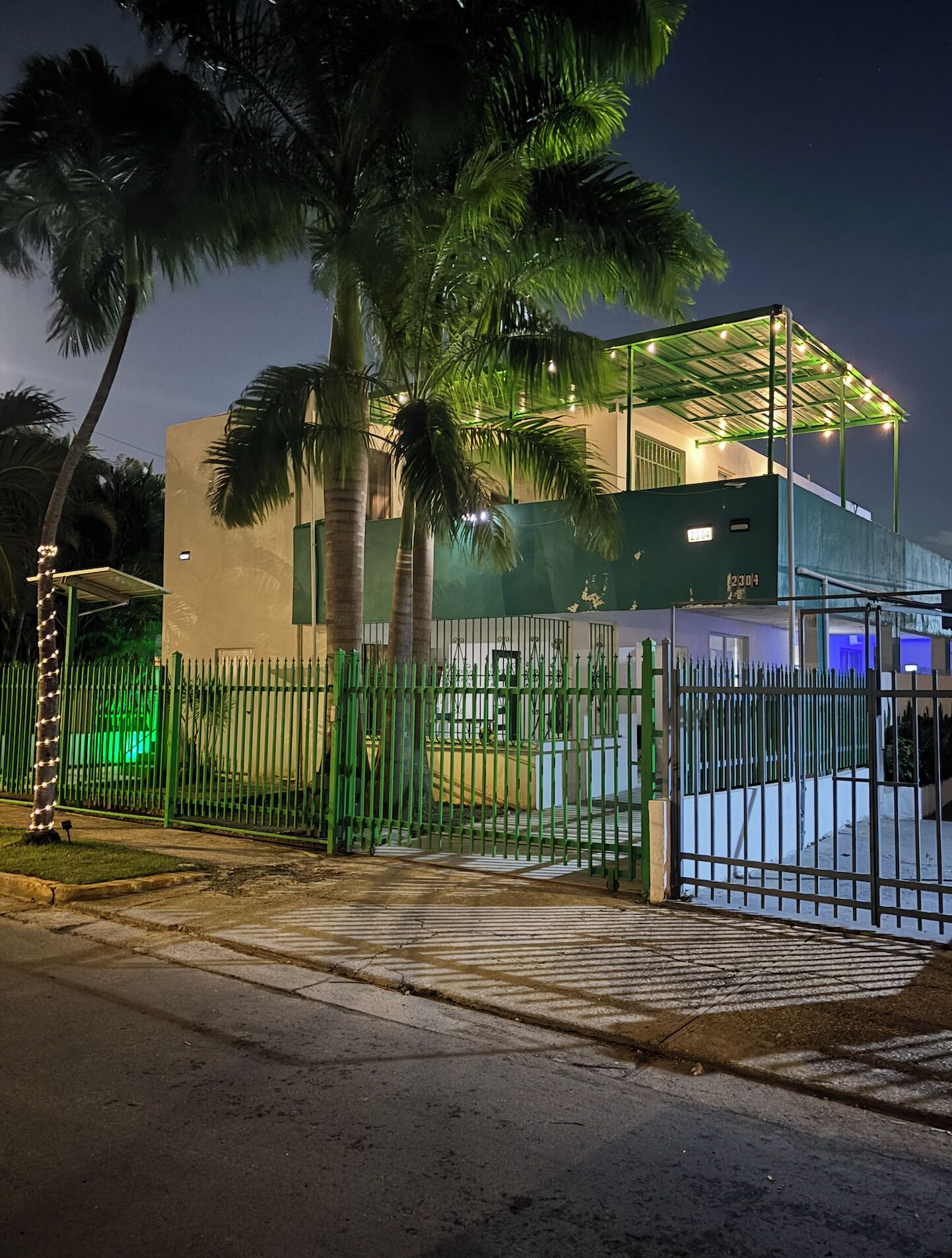Una propiedad cercada con un espacio exterior cubierto, palmeras y un edificio de techo plano.