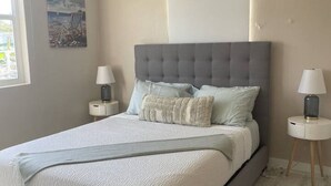 A modern bedroom with a grey upholstered bed, two bedside tables with lamps, a ceiling fan, and a beach-themed painting on the wall. - 3 Bedroom/2 Bathroom 1 block from the beach (San Juan)