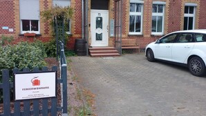 En byggnad av tegel med en vit dörr märkt med siffran ’75’, en bänk och en vit bil parkerad framför.
